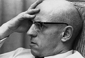 A picture of Michel Foucault, mostly head shot, with his hand on his forehead, wearing glasses, apparently deep in contemplation.